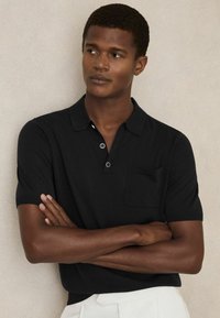 Polo de culoare neagră, cu mâneci scurte și o textură moale, având un guler, placket cu trei butoane și un buzunar pe piept. Styiling cu brațele încrucișate.