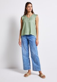 Hellgrünes ärmelloses Oberteil mit V-Ausschnitt und gerafften Schultern, kombiniert mit hellblauen weit geschnittenen Jeans und beigen flachen Sandalen.