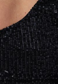 Černá flitrová tkanina s texturovaným povrchem, obsahující překrývající se kulaté flitry, které odrážejí světlo a vytvářejí třpytivý efekt.