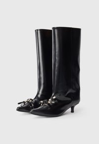 Botas negras hasta la rodilla, hechas de material brillante, con punta afilada, tacón delgado y adornos decorativos en el tobillo.