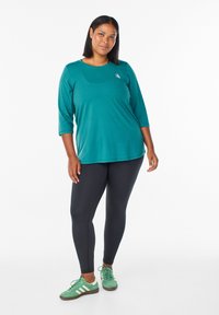 Tyrkysový, dlhý rukáv top s okrúhlym výstrihom, v kombinácii s čiernymi legínami a zelenými teniskami s bielymi akcentmi. Mäkko, elastický materiál.