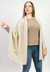 Beige sjal lavet af let stof, med et draperet design og åben front. Båret over en matchende beige top og blå jeans.