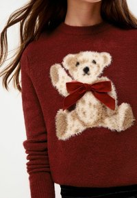 Sarkans adīts džemperis ar pūkainu bēšas krāsas teddy lāča aplikāciju un sarkanu satīna loku ap kaklu.