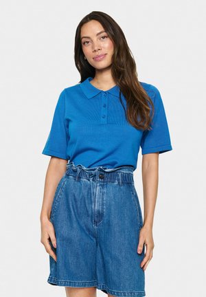 Chemise polo bleue avec un plastron à boutons et des manches courtes, associée à un short en denim bleu clair à taille haute et ceinture froncée.