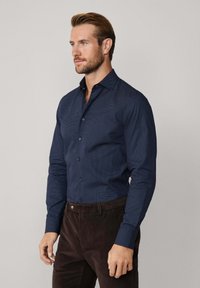 Man met kort haar en baard, gekleed in een marineblauw gestreept overhemd met knopen en donkere bruine broek, staande tegen een simpele achtergrond.