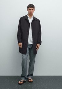 Sort letvægts trenchcoat med store knapper, båret over en hvid skjorte og blå jeans. Modellen står på en struktureret mørk overflade.