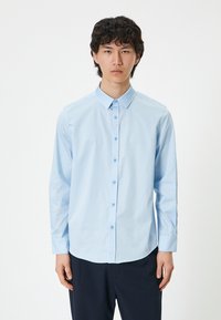 Ljusblå skjorta med knappar, tillverkad av slätt tyg, med krage, långa ärmar och rundad nederkant, kombinerad med mörka byxor.