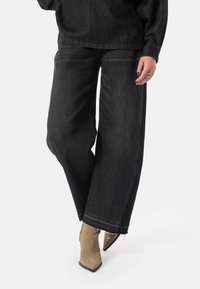 Svarte denim-bukser med vide ben og frynsete kant, kombinert med brune, spisse ankelstøvler. Stoffet har en glatt tekstur og myk finish.