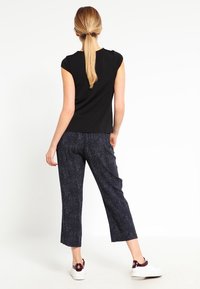 Kobieta stojąca tyłem do aparatu, ubrana w czarną koszulę z krótkim rękawem, ciemne wzorzyste spodenki do kostek oraz białe trampki z bordowymi akcentami.