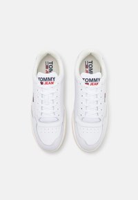 Para białych sneakersów Tommy Jeans z sznurowadłami, czerwonym i granatowym logo na języku i boku, widok z góry na białym tle.