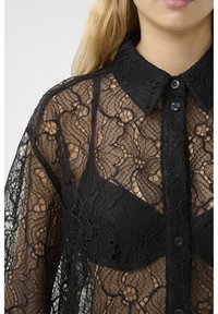 La camicia in pizzo nero presenta motivi floreali, una chiusura con bottoni e un colletto. Sotto, c'è un reggiseno nero abbinato.