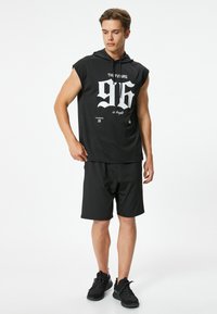 Schwarze ärmellose Kapuzenjacke mit weißem Grafikdruck "96" und Text, kombiniert mit schwarzen Shorts. Der Stoff wirkt glatt und sportlich im Stil.