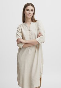 Robe en lin beige avec un col arrondi, des manches trois-quarts et des détails de boutons. Elle présente des fentes sur les côtés pour plus de liberté de mouvement et de confort.
