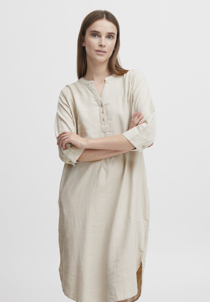 Robe en lin beige avec un col arrondi, des manches trois-quarts et des détails de boutons. Elle présente des fentes sur les côtés pour plus de liberté de mouvement et de confort.