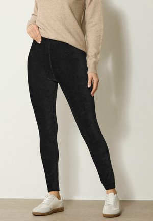 Mustat corduroy-legginssit, joissa on sileä pinta, korkea vyötärö ja tiukka malli, yhdistettynä valkoisiin tennareihin neutraalilla taustalla.