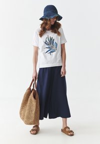 T-shirt blanc avec un motif de feuilles vertes et bleues, culottes larges bleu marine, sandales marron, et sac tissé beige avec des poignées marron. Chapeau bleu.
