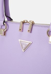 Sac à main synthétique violet avec un logo triangulaire, quincaillerie dorée, surface texturée et double fermeture éclair avec tirettes décoratives.