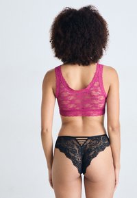 Fuchsiový čipkový bralet s vlnitými okrajmi, skombinovaný s čiernym čipkovým tangami s designom s popruhmi na chrbte. Hladké textúry a jemné vzory.