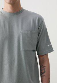 T-shirt gris à manches courtes en coton, doté d'une poche poitrine appliquée et d'un détail logo sur la manche. Texture lisse, design à col rond.