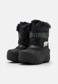 Botas de invierno negras con suelas de goma texturizadas, forro de piel de oveja, correa ajustable y un logo de oso blanco en el costado. Material sintético duradero.