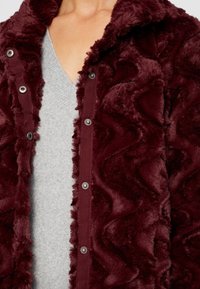 Burgundowa, pluszowa kurtka o falistym wzorze, z zatrzaskami z przodu i miękkiego materiału, noszona na jasnoszarej koszuli.