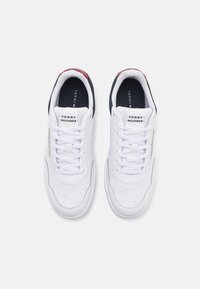 Baskets blanches avec un bout perforé, lacets plats blancs, et une marque sur la languette. Accents en marine et rouge au niveau du talon. Matériau lisse.