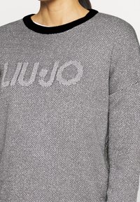 Harmaa teksturoitu neulepusero, jossa musta pyöreä kaula-aukko, rintakehällä vaaleasti neulottua tekstiä.