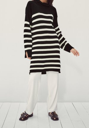 Zwart-wit gestreepte gebreide jurk met lange mouwen, colkraag en zijsplitten. Gecombineerd met losse witte broeken en bruine sandalen.