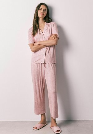 Vrouw staat met armen over elkaar gekleed in een lichtroze kortemouwig pyjamapak met knopen en bijpassende slippers tegen een effen muur.