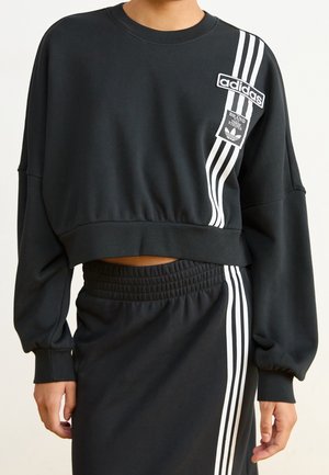 Sweat-shirt court noir Adidas et jupe assortie avec le marquage à trois bandes blanches sur le côté gauche et les logos sur la poitrine et la jupe.