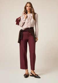 Blusa rosa chiaro con volant, pantaloni tailored bordeaux e ballerine nere con un accento decorativo. Maglione marrone legato in vita. Borsa rossa.