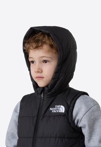 Sort puffervest med hette, glidelåslukking, sydd paneler og en hvit logo på venstre bryst, båret over en grå langermet skjorte.