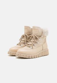 Botas de senderismo beige con parte superior de ante y cuero, cuello forrado de piel, diseño de cordones y suela gruesa con relieve. Presenta ojales metálicos y branding.