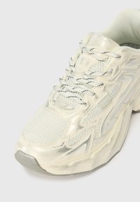 Könnyű sportcipő off-white hálós anyagból és fényes szintetikus anyagokból. Texturált felületi részletekkel és kontrasztos fekete fűzőkkel rendelkezik.