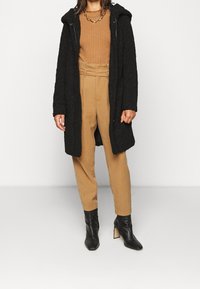 Svart texturerad lång kappa med huva, bärs över en ribbad brun tröja och beige högmidjade byxor. Svarta ankelstövlar fullbordar outfiten.