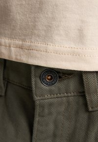 Crème katoen t-shirt met een zoom boven de olijfgroene broek, voorzien van een metalen knop met gegraveerde tekst en een bruine acccent. Zichtbare stiksels.