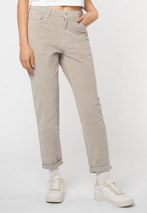 Persoon die beige corduroy broeken met omslag draagt, witte sokken, witte sneakers en een wit cropped topje, staand tegen een effen lichte achtergrond.