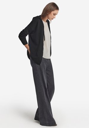 Svart huva cardigan över en ljus topp, kombinerat med vida grå denimbyxor. Outfiten har en avslappnad passform och mjuka texturer.