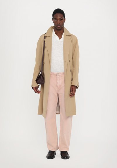 Beige trenchcoat yli harmaan neuleen, yhdistetty vaaleanpunaisiin leveälahkeisiin housuihin ja mustiin kenkiin. Pitelee pientä ruskeaa laukkua.
