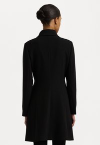Musta takki, joka on valmistettu sileästä kankaasta, pitkässä ja rakenteellisessa muotoilussa, jossa on viisto kaulus ja sauma-aronkompi. Elegantti ja virtaviivainen muoto.
