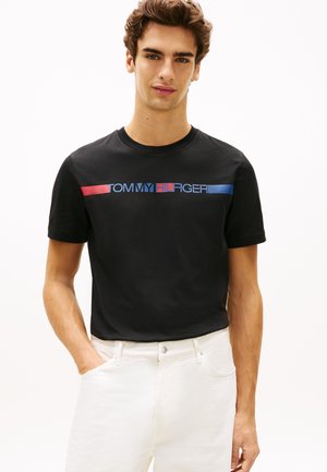 Mladić s kratkom smeđom kosom, nosi crnu Tommy Hilfiger majicu i bijele hlače, stojeći s jednom rukom u džepu.