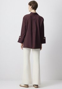 Camicia marrone con colletto, maniche lunghe con bottoni dorati, abbinata a pantaloni bianchi svasati e scarpe beige. Design semplice e pulito.