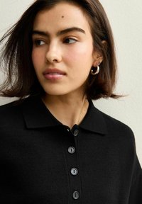 Zwarte gebreide poloshirt met een kraag, voorzien van vijf zwarte knopen aan de voorkant. Soepele textuur met korte mouwen. Eenvoudig ontwerp zonder patronen.
