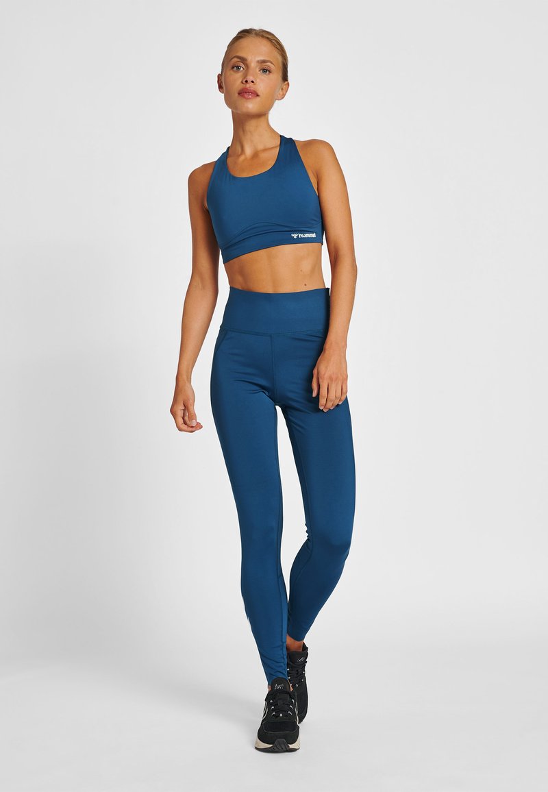 Kvinna i blå sport-BH och högmidjade leggings poserar mot en vit bakgrund, bärande svarta träningsskor.