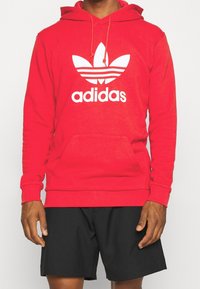 Roter Adidas-Hoodie mit großem weißem Logo und drei Streifen-Design, mit einer Fronttasche und verstellbarem Kordelzug für die Kapuze. Kombiniert mit schwarzen Shorts.