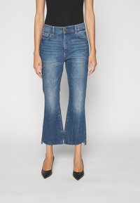 Jeans de mezclilla azul cortos con un diseño acampanado, dobladillo sin rematar, bolsillos delanteros y cierre de botón, combinados con tacones altos negros.