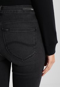 Jeans de denim negro con un diseño ajustado, que presentan una costura curva distintiva en los bolsillos traseros y una etiqueta de marca sutil.
