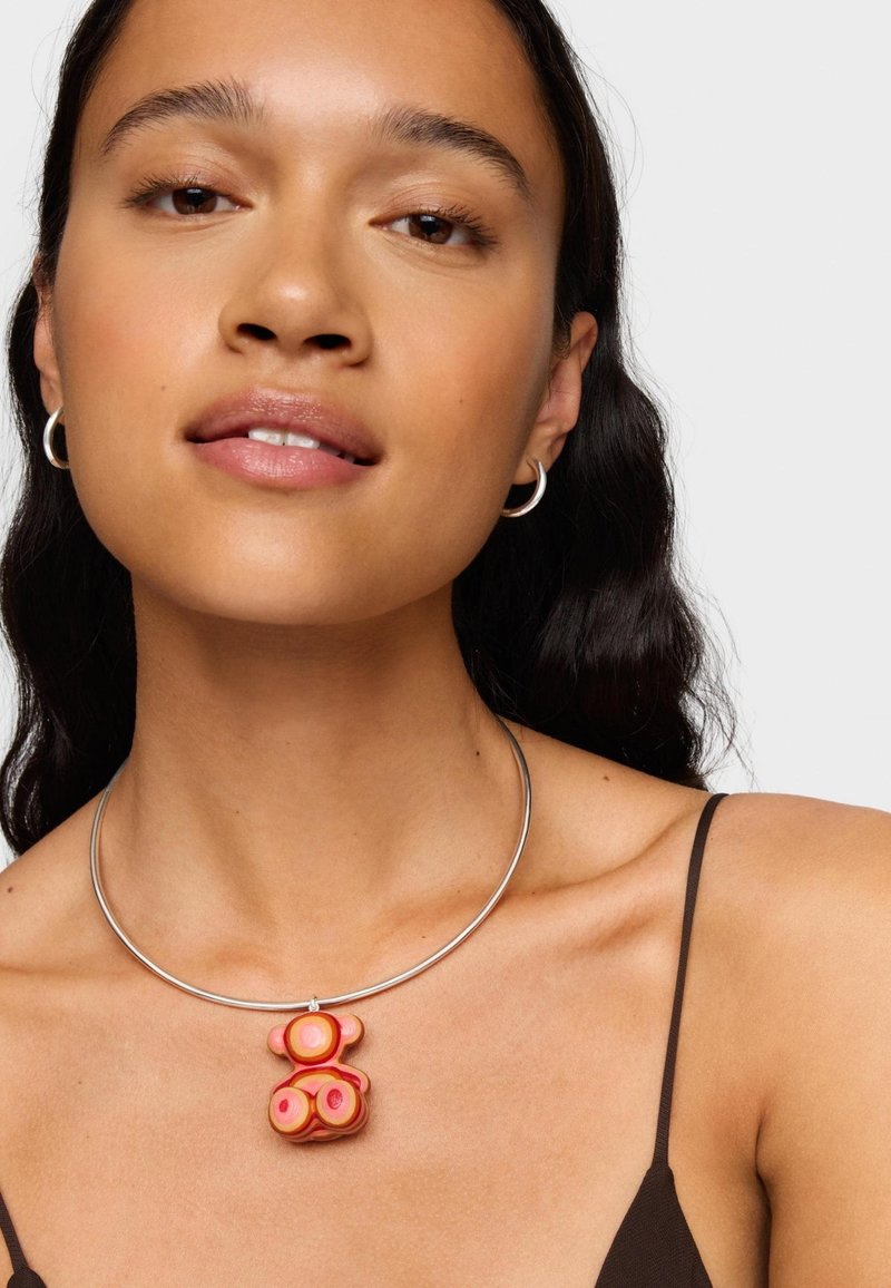 Ciondolo colorato a forma di orso in tonalità rosa su una collana d'argento, texture liscia, design circolare, indossato con una canotta scura.