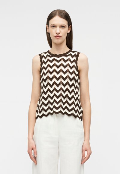 Γυναίκα που φοράει αμάνικο πλεκτό τοπ με καφέ και λευκό σχέδιο ζιγκ-ζαγκ και ψηλόμεσο λευκό παντελόνι, στέκεται μπροστά σε απλό φόντο.
