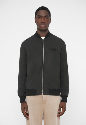 Sort bomberjakke i letvægtsstof med ribstrikket krave og manchetter, lynlåslukning foran, brystmærkat, kombineret med lysebeige bukser.